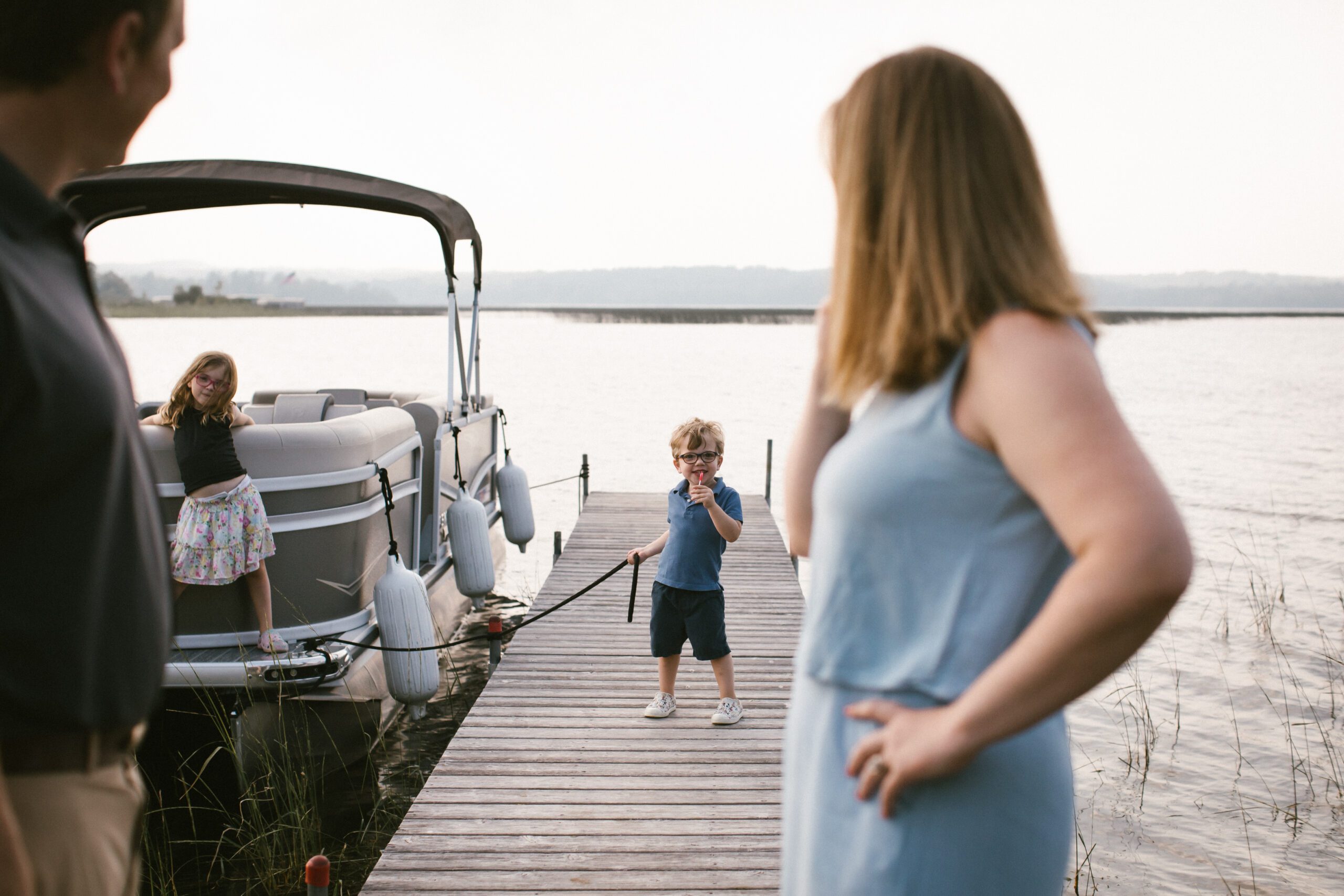 A family photography session in Minnesota on a lake, candid, with parents watching kids be silly on the dock near the boat at sunset
